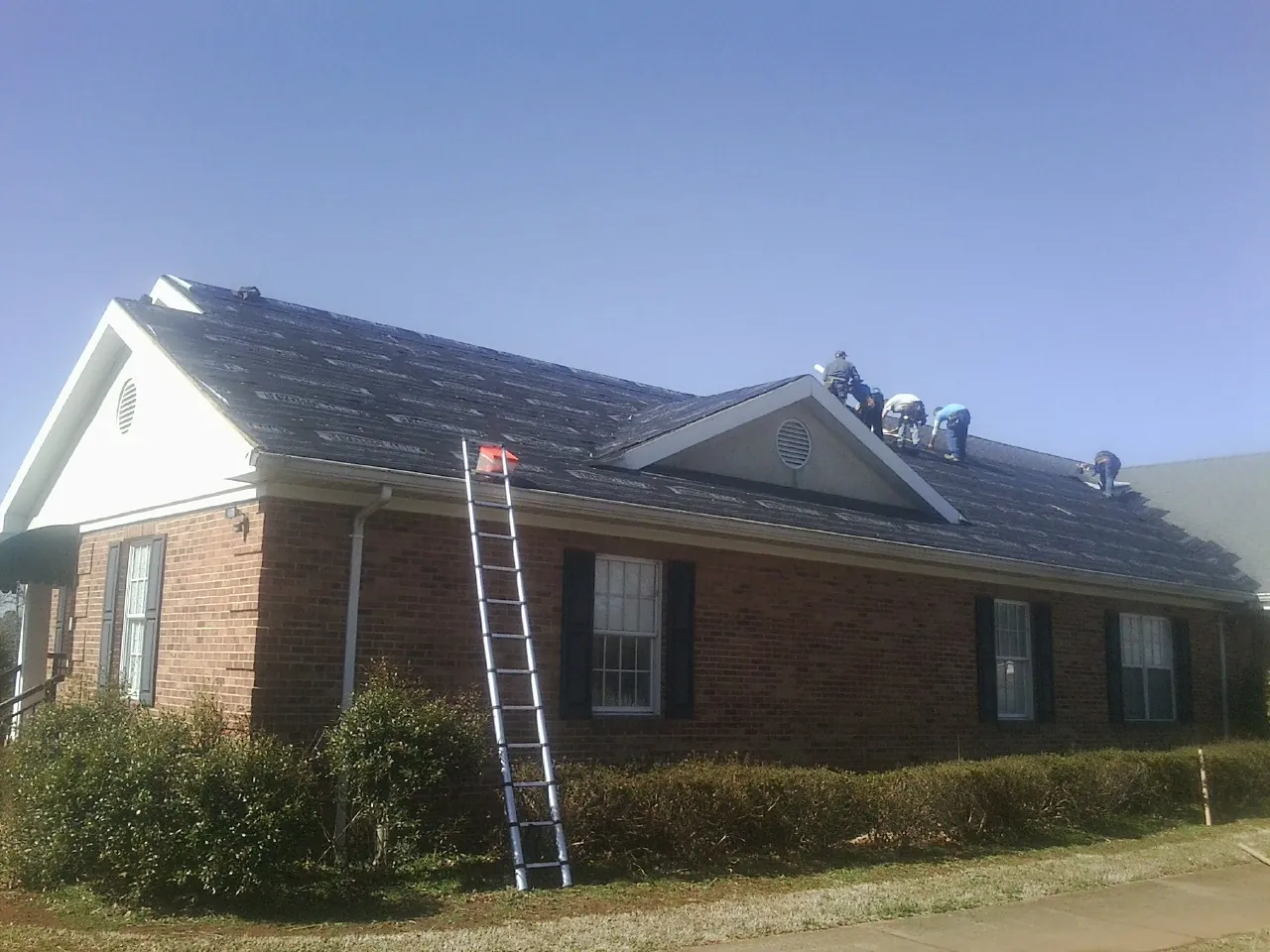Crew tearing shingles off a brick ranch roof with a ladder set — Metal Roof Repair work by our crew in Harmony